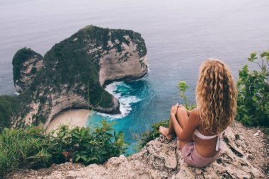Mayo giymiş isimsiz kadın turistin kayalık kayalıklarda oturup tatilin tadını çıkarırken kumsalı ve tepesi olan engin mavi deniz manzarası.