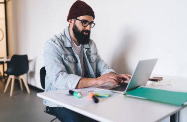 Konsantre sakallı, günlük giysiler ve gözlüklerle masada oturmuş dizüstü bilgisayara bakarken not defteri yapıştırıyor ve modern çalışma alanında yeni bir başlangıç için çalışıyor.