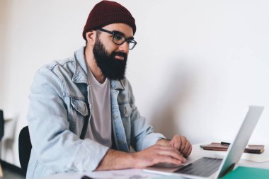 Gündelik giysiler ve gözlük takan sakallı bir erkek, dizüstü bilgisayara bakarken elinde kalemle masada oturuyor ve masa başı işlerde çalışıyor.