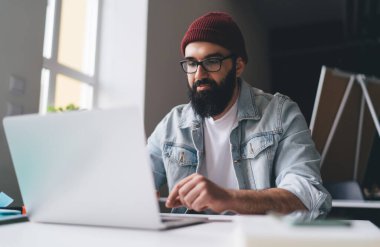 Akıllı giyimli, gözlüklü, dizüstü bilgisayarda gezen ve modern yaratıcı çalışma alanında iş projesinde çalışan konsantre sakallı serbest bir erkek.