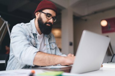 Sakallı, serbest çalışan, şık giyinen, netbook 'la masaya oturan ve yaratıcı ofiste proje üzerinde çalışan bir erkek.