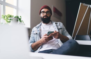 Kot ceketli ve şapkalı ciddi sakallı bir erkek işçi masada oturmuş kameraya bakarken akıllı telefon kullanıyor.