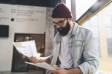 Düşük açılı, gözlüklü bir erkeğin güneş ışığının yanında otururken elindeki belgeleri tek tek karıştırması ofis girişinin dışındaki cam yansıtması.