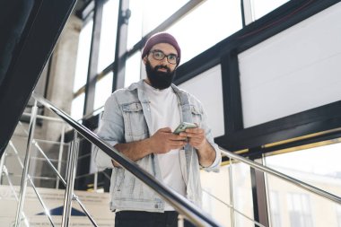 Kot ceketli, şapkalı, cep telefonlu genç sakallı hippi merdivende büyük pencerelerin yanında kameraya bakıyor.