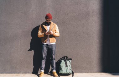 Sokak tarzı kıyafetli sakallı bir erkek hippi, cep telefonu web sitesi kullanırken sırt çantasıyla güneş ışığına karşı ayakta duruyor.