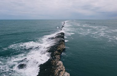 Düz olmayan yüzeyi ve köpüklü dalgaları olan engebeli kayalık manzarası gündüz vakti alçak irtifada bulutlu gökyüzüne karşı geçerken su sıçrıyor ve kırılıyor.