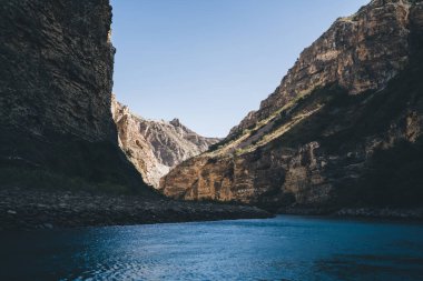 Dengesiz yüzeyi olan engebeli kayalık dağların manzarası. Gün ışığında mavi suyun gölgesi ve bulutsuz gökyüzü arasında akması.