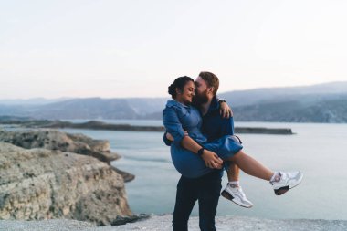 Denize yakın çok ırklı bir çiftin gözleri kapalı kayalık kayalıklarda dikilirken ve mutlu vakit geçirirken erkeklerin elleri karışık bir ırkın alnında nazikçe öpüşmeleri ve kucaklaşmaları.