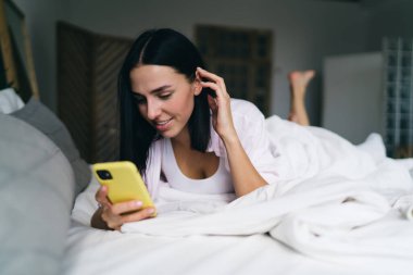 Konsantre olmuş genç bir kadın cep telefonuyla rahat bir yatakta uzanıyor ve yatak odasında dinlenip sohbet ederken ekrana bakıyor.