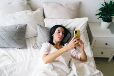 Yüksek açılı, gülümseyen genç bir kadın ekrandaki cep telefonuna bakıyor ve yatakta sohbet ediyor.