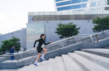 Modern şehir merkezinde siyah giysili genç bir bayanın yan görüntüsü ve cam binalara karşı beton merdiven çıkıyor.