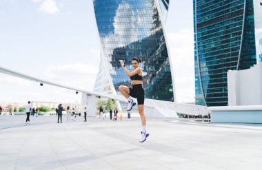 Spor kıyafetleri ve spor ayakkabıları giyen formda bir bayan sporcunun modern binaların yakınındaki şehir caddesinde koşu egzersizi yaparken gündüz kameralarına bakması.