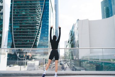 Siyah kıyafetli, camdan balkonda duran, kollarını kaldırmış ve gündüzleri şehrin manzarasına hayran olan anonim bir kadının arka görüntüsü.