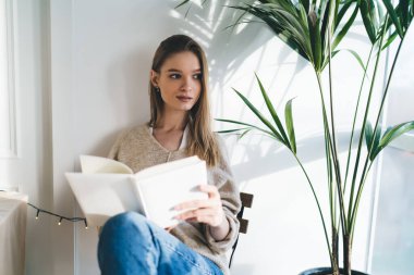 Evde kompozisyon yazarken ve uzaktan öğrenirken pencerenin yanında oturan günlük kıyafetli pozitif bayan öğrenci.