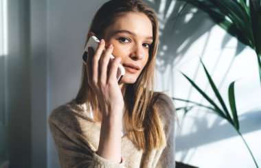 Günlük kıyafetlerle gülümseyen genç bir kadın kameraya bakarken yeşil bitkinin yanında otururken beyaz duvarda güneş tonu ve cep telefonuyla konuşuyor.