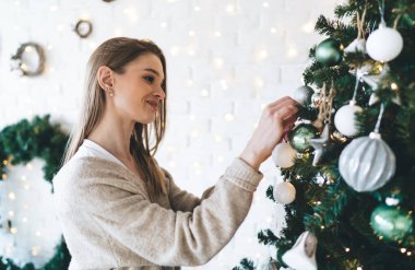 Yeni yıl kutlamalarına hazırlanırken sıcak süveter giyen, gülümseyen ve Noel ağacını süsleyen neşeli genç bir kadın.