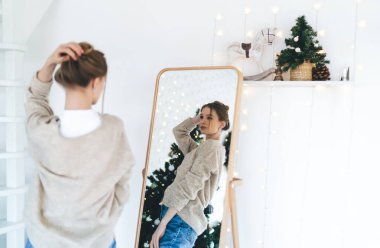 Yılbaşı ağacı süsleyerek evde vakit geçirirken aynaya bakıp saçlarına dokunan gündelik elbiseli genç bir kadının arka plan görüntüsü.