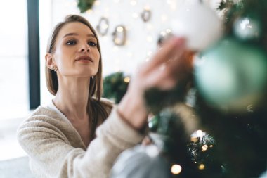 Kış tatilinde Noel kutlaması için apartman dekorasyonu yaparken sıcak süveterli sakin bir kadın Noel ağacına takılıyor.