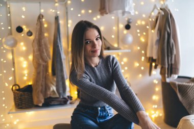 Parlayan ışıklara karşı otururken ve butikte kıyafetleri asarken gülümseyen ve kameraya bakan sıradan bir kadın.