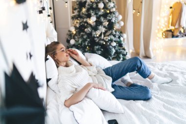 Yan bakış açısı beyaz kazaklı genç bir kadın dekore edilmiş yatak odasında rahat bir yatakta dinleniyor gözlükleri ayarlıyor.