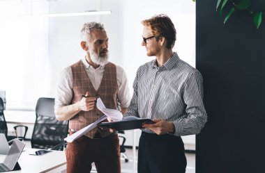 Üst düzey sakallı girişimci modern ofiste duruyor ve iş günlerinde genç bir erkek arkadaşla iş konularını tartışıyor.