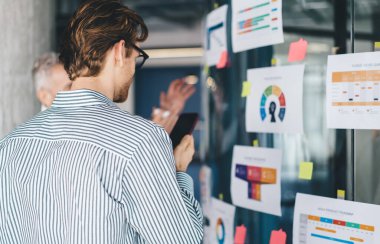 Çizgili gömlekli ve gözlüklü genç bir erkek çalışanın iş sırasında cam duvarın yanında çıkartma ve diyagramlarla dururken cep telefonu kullanışının arka planı