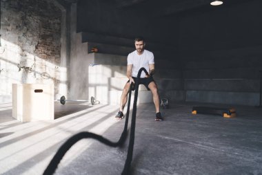 Güçlü erkek sporcuların yan görünüşü peş peşe halat dövüşü omuzlarda dayanıklılık oluşturmak için spor salonunda ayakta ve pratik yaparken.