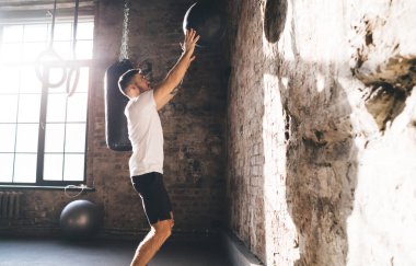 Beyaz tişörtlü kararlı sporcunun spor salonundaki kardiyo eğitimi sırasında duvara top atması.