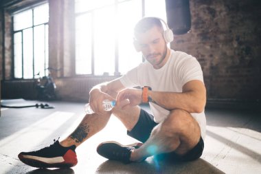 Kulaklık ve aktif giyimli arka ışıklandırmalı beyaz erkek atlet modern ferah fitness stüdyosunda yerde bir şişe suyla otururken akıllı kol saati ile nabız ölçümü yapıyor.