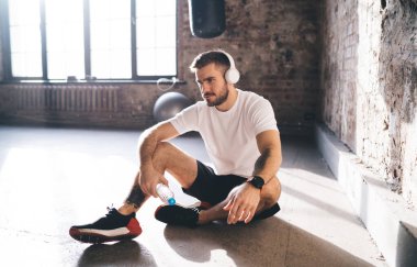 Spor giyim sektöründe sakallı bir erkek sporcu spor yaparken dinleniyor ve antrenman sırasında kulaklıkla müzik dinliyor. O sırada da eski püskü spor salonunda yerde bir şişe suyla oturuyor.