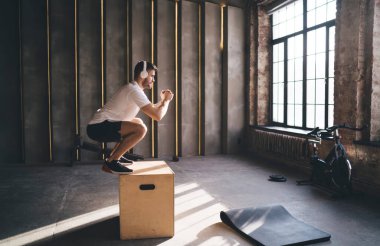 Aktif giyim ve kulaklık giyen sportif erkek sporcu atlama kutusu üzerinde egzersiz yapıyor ve modern çatı katı tarzı stüdyoda işlevsel eğitim sırasında müzik dinliyor.