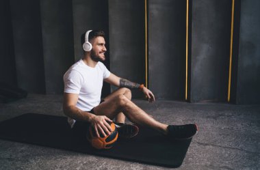 Minderde oturan ve molada dinlenirken kablosuz kulaklıkla müzik dinleyen gülümseyen ince adamın yan görüntüsü