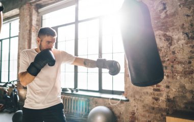 Erkek boksörün boks eldivenleriyle kum torbasına vurması ve güneşli spor salonunda antrenman yapması bir sonraki müsabakayı yerel düzeyde kazanmaya kararlı.
