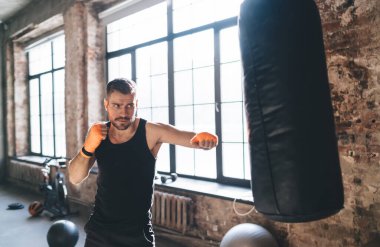 Güçlü kararlı erkek boksör ellerini bantla sarmalamış ve öndeki kum torbasına ekipmanla saldırmaya hazırlanırken gözünü kaçırıyor.