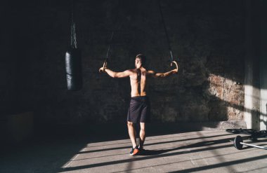 Güçlü kaslı erkek sporcunun tüm vücut arkası görüntüsü kum torbasının yanında sarmalanır ve spor salonunda egzersiz yaparken jimnastik yüzükleriyle antrenman yapmaya hazırlanır.