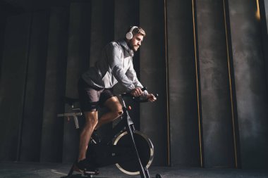 Spor kıyafetleri içinde sakallı bir adamın spor salonunda spor yaparken bisiklet simülatörü üzerinde egzersiz yapması ve spor salonunda egzersiz yaparken kablosuz kulaklıkla müzik dinlemesi.