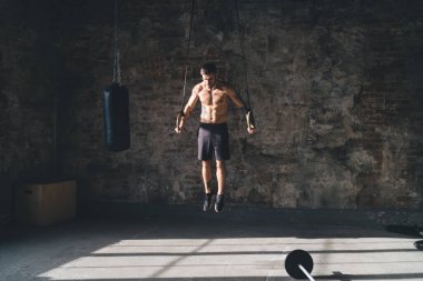 Geniş güneşli stüdyoda kardiyo eğitimi sırasında spor kıyafetleriyle jimnastik halkaları üzerinde kompleks bir duruş sergileyen üstsüz ve güçlü etnik erkek sporcu.