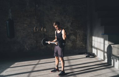 Çağdaş spor kulübünde ağırlık kaldırma egzersizi sırasında ağır halter kaldıran kararlı erkek sporcunun tüm vücudu güneş ışığıyla pencerenin önünde duruyor.