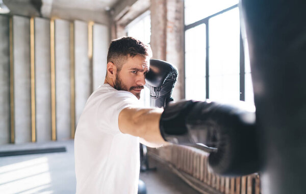 Вид сбоку на бородатого спортсмена в белой рубашке и боксерских перчатках, бьющего боксерскую грушу во время тренировки в освещенном солнцем спортзале