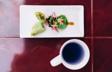 Masanın üzerinde beyaz bir fincan sıcak mavi içecek, taze kesilmiş meyveler ve sosla süslenmiş lezzetli sebzeler. Masaya seramik bir tabakla süslenmiş çiçekler.