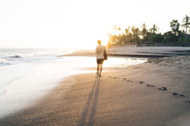 Gün doğumunda Bali 'de parlak güneş ışığı altında el çantalı, kimliği belirsiz bir adamın el çantalı, ıslak kumlu sahilde el sallayarak, palmiye ağaçlarıyla tatil köyüne doğru yürüyüşü.