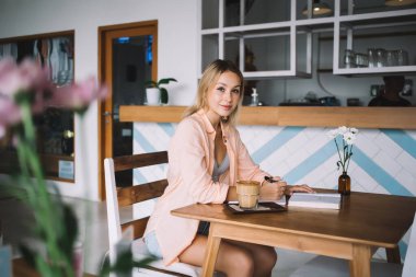 Bir yandan gülümseyen genç bir kadının elinde bir bardak kahveyle masaya otururken ve deftere kalemle ön planda bulanıklık olan bir kitaptan notlar alırken kameraya bakması.