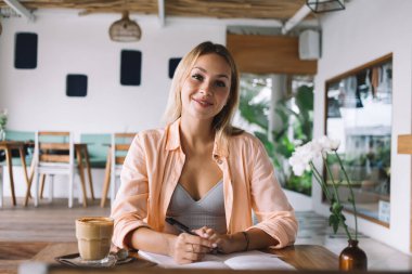 Bir yandan gülümseyen ve kameraya bakan genç bir kadın, elinde bir bardak kahve defteri ve kalemle kafede otururken, ayna ve bulanık bir yansımayla.