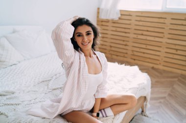 Uzun siyah saçlara dokunan ve güneşli bir sabahta rahat bir yatakta otururken kameraya gülümseyen kendine güvenen genç İspanyol kadın milenyumu.