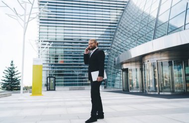 Siyah takım elbiseli sakallı bir işadamının modern ofis binasının yanında cam duvarlarla konuşurken cep telefonuyla konuşmasının yan görüntüsü.