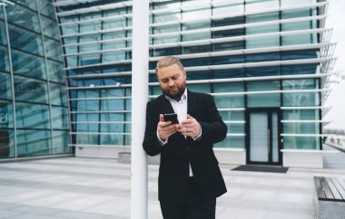 Gündüz vakti modern cam duvar binasının girişinde dikilirken ekrana bakarken ve beyaz direğe yaslanırken konsantre erkek girişimci cep telefonuna göz atıyor.