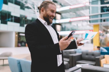 Resmi olarak iyimser erkek girişimcinin yandan görünüşü, modern ofis salonundaki akıllı telefonları kontrol ederken gülümseyen ve kağıt dökümanlarla ayakta duran giyim tarzı.