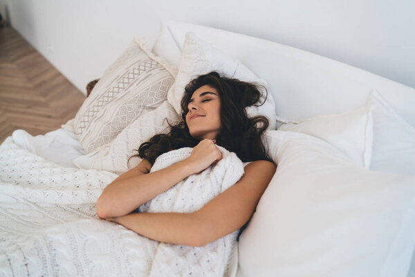 Сверху положительная молодая женщина лежит под теплым одеялом во время сна на удобной кровати с закрытыми глазами у себя дома