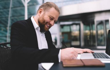 Pozitif sakallı, klas takım elbiseli serbest çalışan, masa başında oturup notlar yazan genç bir erkeğin yan görüntüsü.