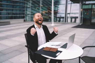 Şık takım elbiseli neşeli, sakallı, uzaktan kumandalı bir erkek bilgisayar başında oturmuş modern şehrin sokaklarına bakarken başarıyı kutluyor.